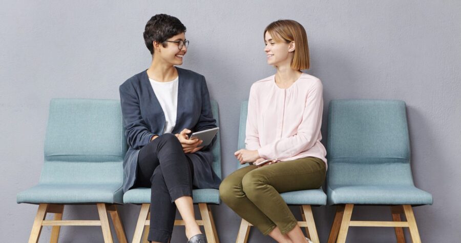 Lively conversation in queue. Two unemployed attractive young women in strict clothing talking to each other at HR department and discussing their resumes while waiting their turn for job interview